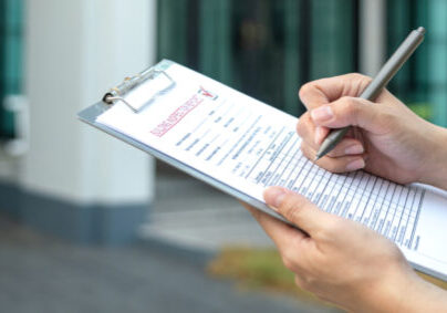 Foreman's hand is using a pen checking on building inspection report form to QC building quality of the house (background). Construction industrial working scene concept. Close-up and selective focus.